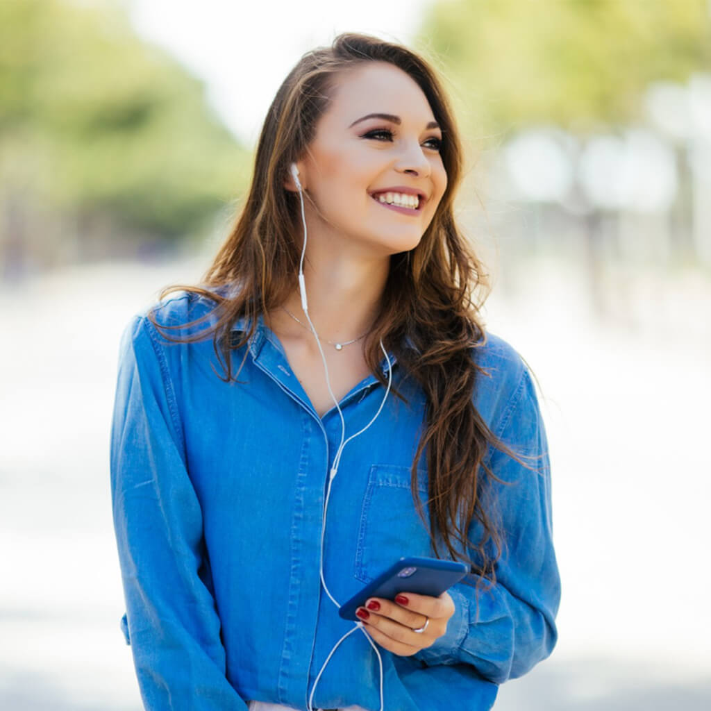 Smiling woman in a blue shirt using her phone and listening to music with earbuds outdoors.