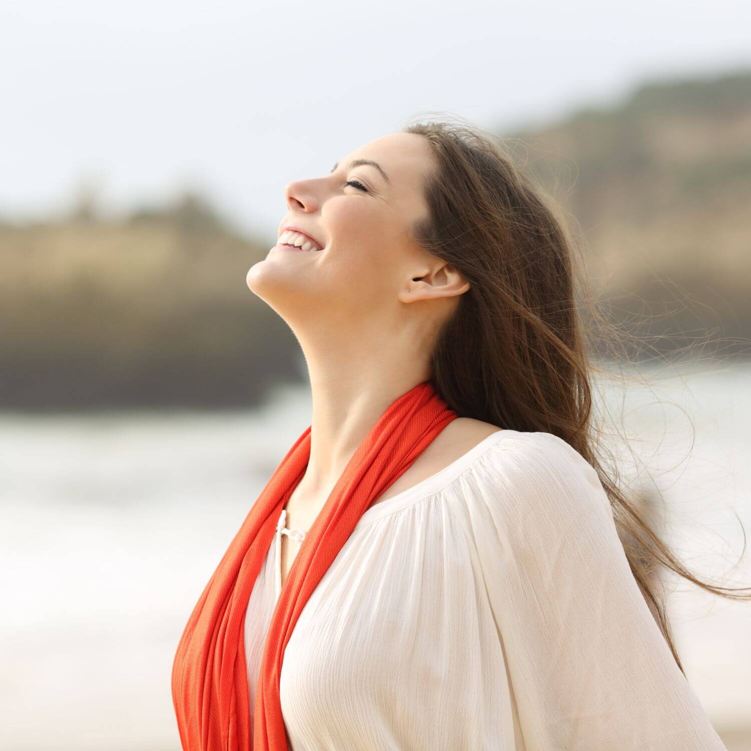 Woman smiling joyfully on the beach wearing a red scarf, embodying a refreshing and carefree spirit.