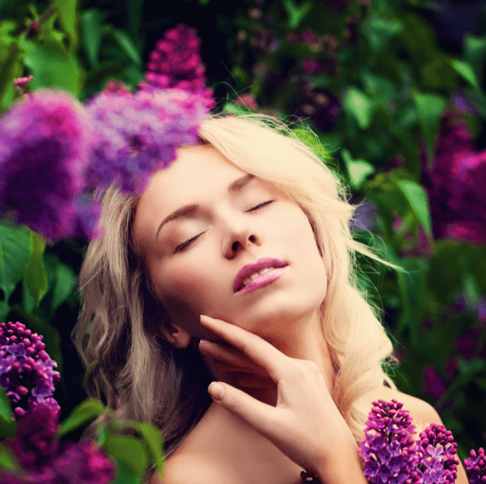 Woman enjoying the beauty of nature surrounded by lilac flowers, representing pampering skincare with Luxe Skin Repair Cream.