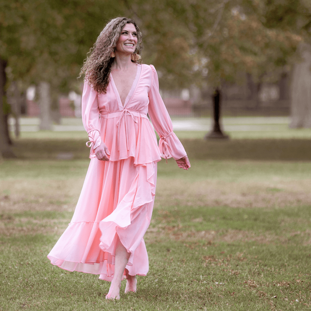 Woman in a flowing pink dress walking barefoot in a park, enjoying a beautiful day outdoors.