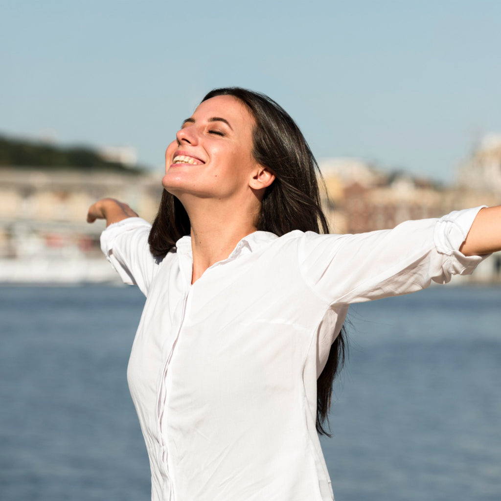 Woman enjoying the outdoors with arms open, expressing joy near water, showcasing happiness and self-care.