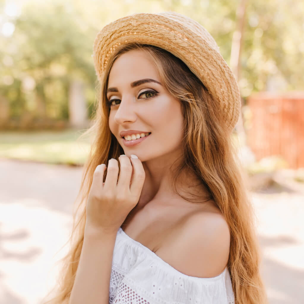 Young woman with long hair wearing a straw hat, smiling and looking radiant outdoors.