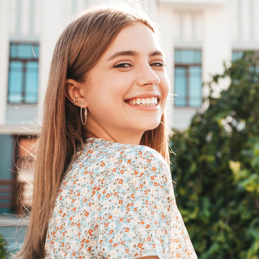 A young woman smiling joyfully while wearing a floral top, enjoying a sunny day outdoors.
